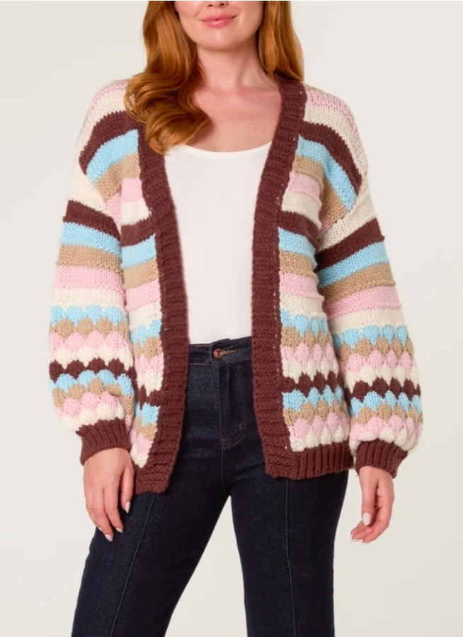 A woman wearing a chunky knit open-front cardigan with horizontal stripes in brown, beige, pink, light blue, and cream, styled with a white top and dark blue jeans against a plain light background.
